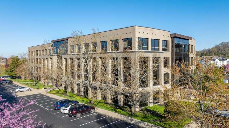 A multi-story office building with large windows and light brick exterior, surrounded by trees and a parking lot.