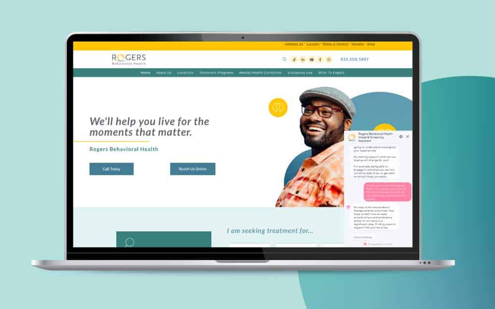 The image showcases a laptop displaying the Rogers Behavioral Health website. The screen presents a modern homepage design featuring a prominent banner with the text, "We’ll help you live for the moments that matter." Below the text are options to call or reach them online. The right side of the screen shows a smiling man wearing glasses and a hat, augmented by circular graphics in blue and yellow.