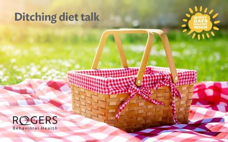 The image portrays a wicker picnic basket with red and white checkered fabric lining and handles made of light wood. It sits on a matching red and white checkered picnic blanket. The scene is set outdoors in a sunny park with a blurred background of green grass and white flowers, suggesting a leisurely atmosphere. In the top left corner is the text "Ditching diet talk," and in the top right corner, there is a sun-like graphic with the words "Sunny Days Healthy Minds."
