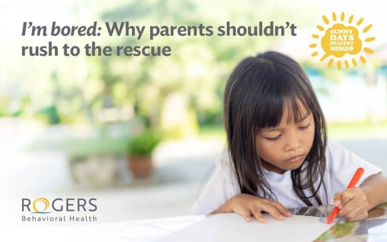 The image shows a young child with long dark hair, wearing a white shirt, concentrating on drawing with a red pencil on a piece of paper. The background is blurred, featuring a bright outdoor setting with green foliage, creating a soft focus. In the top left corner, there is text in bold and regular fonts. In the top right corner, there is a sun graphic with text inside. : Sunny Days, Healthy Minds. Transcribed Text:I’m bored: Why parents shouldn’t rush to the rescue "I’m bored: Why parents shouldn’t rush to the rescue