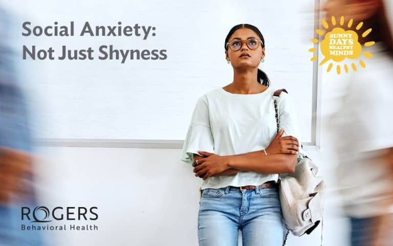 The image shows a young woman standing against a light-colored wall, appearing contemplative. She has her arms crossed and is holding a light gray bag with a brown strap over her shoulder. She wears glasses, a light mint-green blouse, and blue jeans. The background features blurred figures moving past her, conveying a sense of busyness or a crowded environment. The top left corner has large text that reads "Social Anxiety: Not Just Shyness." In the top right corner, there's a sun-shaped emblem with the words "Sunny Days Healthy Minds."