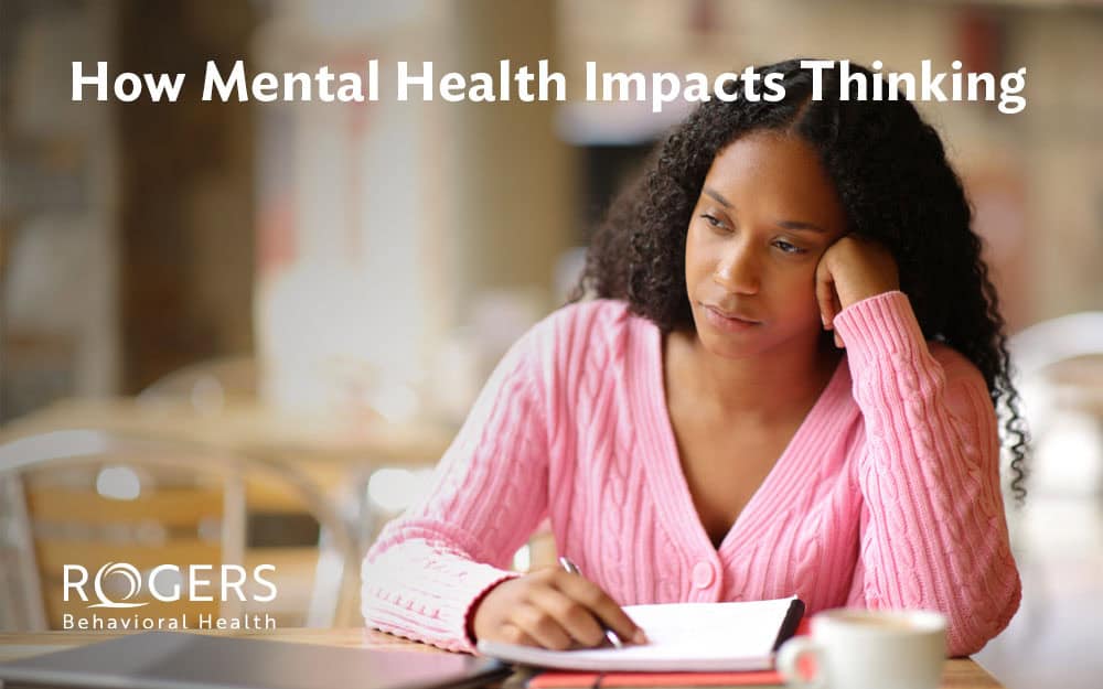 The image depicts a person with a contemplative expression, sitting indoors at a table. They are wearing a pink cable-knit sweater and have long, curly hair. Their chin rests on their hand, while the other hand holds a pen above an open notebook. The background is softly blurred, suggesting a cozy café setting with warm, neutral tones. Transcribed Text: How Mental Health Impacts Thinking