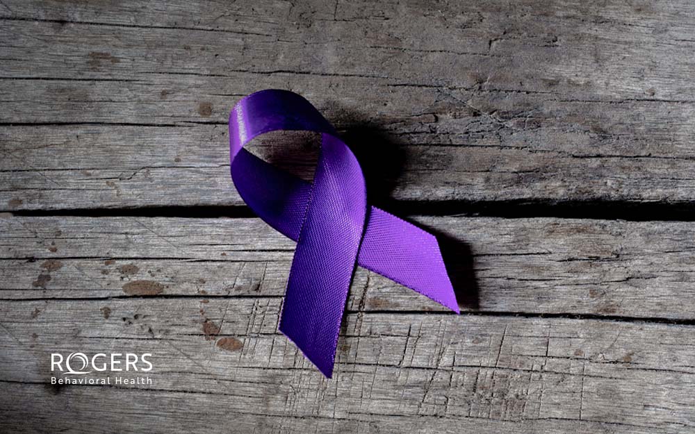 The image shows a purple awareness ribbon resting on a weathered wooden surface with visible grain and texture. The ribbon forms a loop with crossed ends, casting a shadow on the wood. The background consists of horizontal wooden planks of varying shades of gray and brown, exhibiting signs of age and wear.