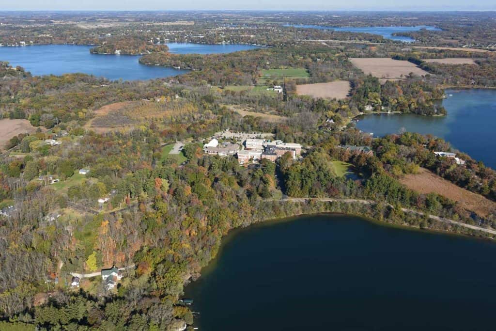 Aerial view of a landscape with water, trees in autumn colors, and a complex of buildings surrounded by fields.