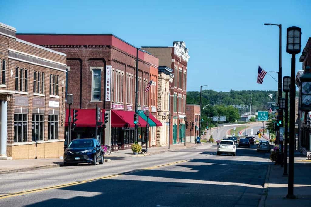 Downtown street scene in Chippewa Falls, Wisconsin, featuring historic brick storefronts, American flags, parked and moving vehicles, and a roadway leading toward a tree-lined area in the distance under a clear blue sky.