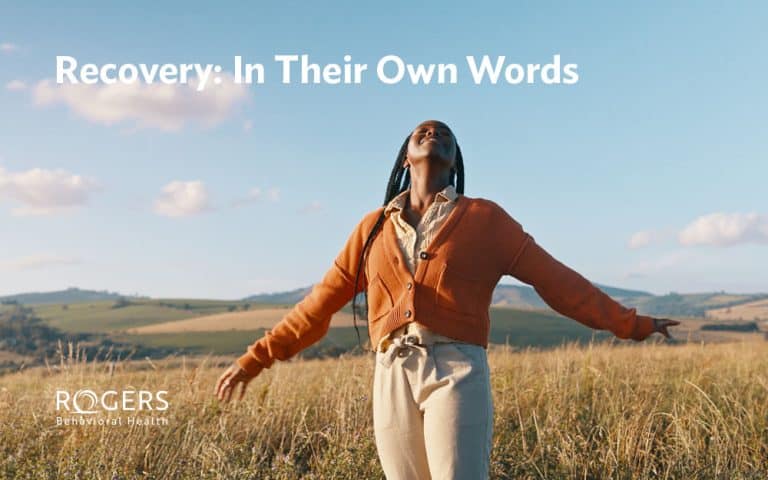 The image depicts a person standing in an open field with tall grass under a clear blue sky. The individual is wearing a bright orange cardigan over a light-colored shirt and beige trousers. They have their arms outstretched, and their face is lifted upwards, suggesting a sense of freedom or joy. In the background are rolling hills covered with patches of grass and distant trees. Transcribed Text: Recovery: In Their Own Words
