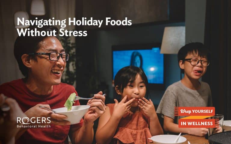 The image shows three people sitting at a dining table, appearing to enjoy a meal together. On the left, a man wearing glasses and a red shirt is smiling while holding a bowl with chopsticks. In the center, a young girl with long hair, wearing a sleeveless top, is touching her lips in a playful manner. On the right, a boy with glasses, wearing a gray shirt, appears to be making a funny face. The background is dimly lit with a large lamp and a blurred television screen visible. Text overlays the image: Navigating Holiday Foods Without Stress Wrap Yourself in Wellness