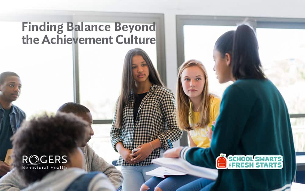 The image depicts a group of five young individuals engaged in a discussion in a well-lit indoor setting. They are seated and standing around a table, which is not visible. The background includes large windows allowing natural light into the room. The focus is on one individual, a young woman with long dark hair, who is speaking while others attentively listen. She wears a teal jacket and holds some papers or a notebook. To her left, another young woman in a yellow shirt and long light brown hair seems attentive and engaged. A young man with short hair and wearing a denim jacket sits close to the viewer, partially visible. Text is prominently displayed at the top : Finding Balance Beyond the Achievement Culture