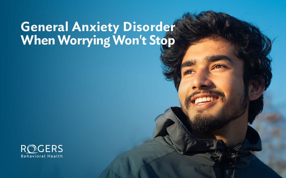 The image features a man with a smile, looking up and to the right. He has dark hair and a beard, and is wearing a gray jacket. The background is a gradient of blue, transitioning from dark to lighter shades, suggesting an outdoor setting. In the upper left corner, white text reads, "General Anxiety Disorder When Worrying Won’t Stop."