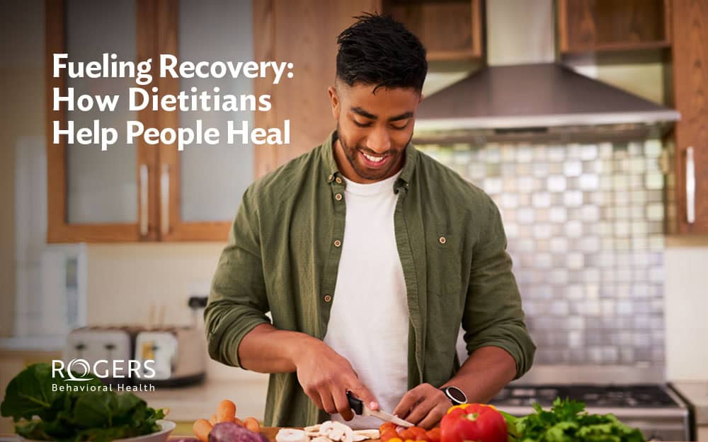 The image depicts a person in a modern kitchen setting, smiling and engaged in slicing vegetables on a wooden cutting board. The kitchen features a metallic backsplash, wooden cabinets, and a stainless steel hood. A variety of fresh produce, including carrots and tomatoes, are visible on the countertop. The person is wearing a green button-up shirt over a white t-shirt. Soft lighting creates a warm and inviting atmosphere. Transcribed Text: Fueling Recovery: How Dietitians Help People Heal