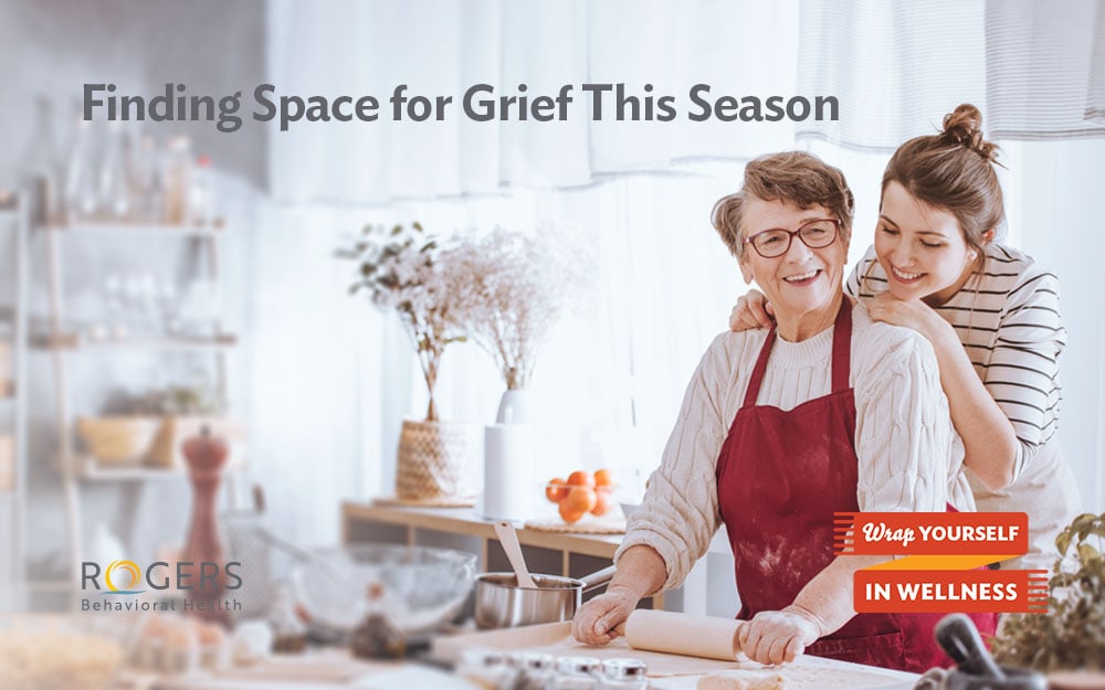 The image features an elderly woman and a younger woman in a kitchen. The elderly woman, wearing glasses, a red apron, and a cream sweater, is using a rolling pin on dough. The younger woman is standing behind her, gently resting her hands on the elderly woman's shoulders, expressing a supportive and joyful demeanor. The background includes shelves with kitchen items and a vase with dried flowers. The setting is warmly lit, creating a cozy atmosphere. Transcribed Text: "Finding Space for Grief This Season. Rogers Behavioral Health. Wrap Yourself in Wellness."