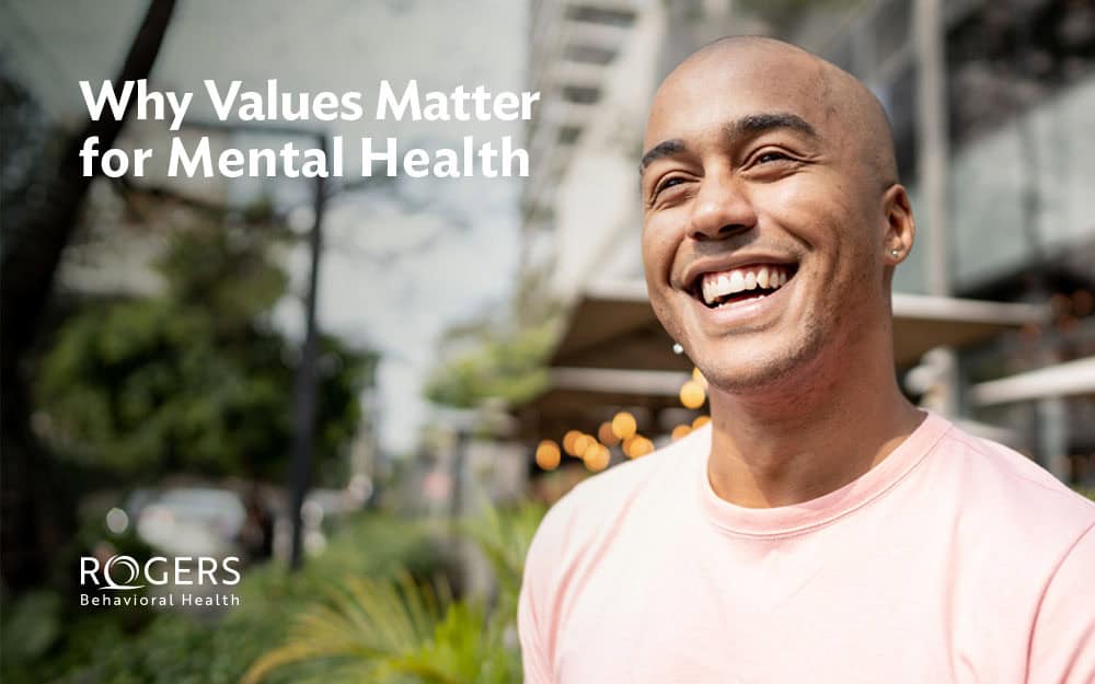 The image features a person standing outdoors, smiling broadly. They are wearing a light pink shirt and have pierced ears. The background includes blurred elements of trees and a building, likely a cafe or restaurant, with small round lights hanging outside. The lighting is bright, indicating a sunny day, and the overall atmosphere is cheerful. The image has text overlaid in the upper left corner. Alt-text: A smiling person in a pink shirt with an outdoor cafe background. Transcribed Text: Why Values Matter for Mental Health