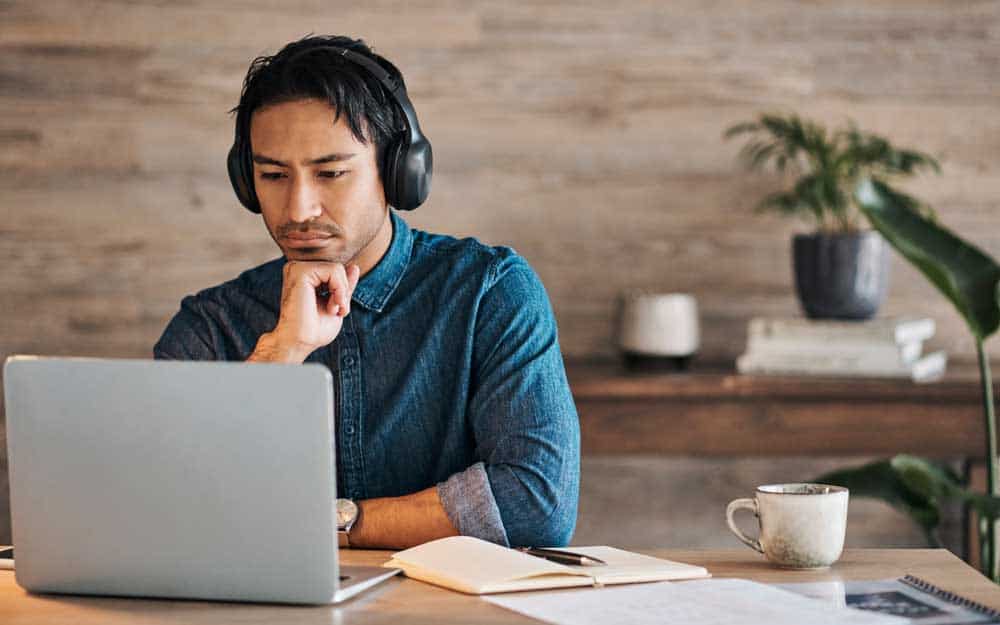 Man wearing headphones, seated at a desk with a laptop and notebook.