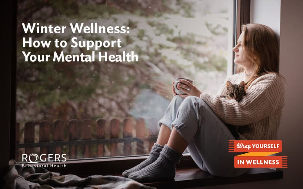 The image features a woman sitting on a window sill, gazing outside. She is wearing a cozy knitted sweater and woolen socks, with her legs pulled up in a relaxed posture. In her arms, she cradles a small cat. Outside the window, there is a blurred view of a snow-dusted landscape with trees and a wooden fence. The scene conveys a sense of warmth and comfort indoors juxtaposed with a cold winter landscape outside. Text is displayed on the left: Winter Wellness: How to Support Your Mental Health ROGERS Behavioral Health Wrap Yourself in Wellness