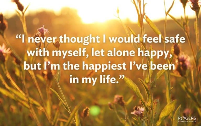 The image features a warm, sunlit field filled with tall grass and wildflowers. The sunlight creates a golden glow over the scene, with the sun setting or rising in the background. The tops of the grasses and flowers are highlighted by the light, casting soft shadows and creating a tranquil atmosphere. The image prominently features a quote written in bold white text across the center: “I never thought I would feel safe with myself, let alone happy, but I’m the happiest I’ve been in my life.”