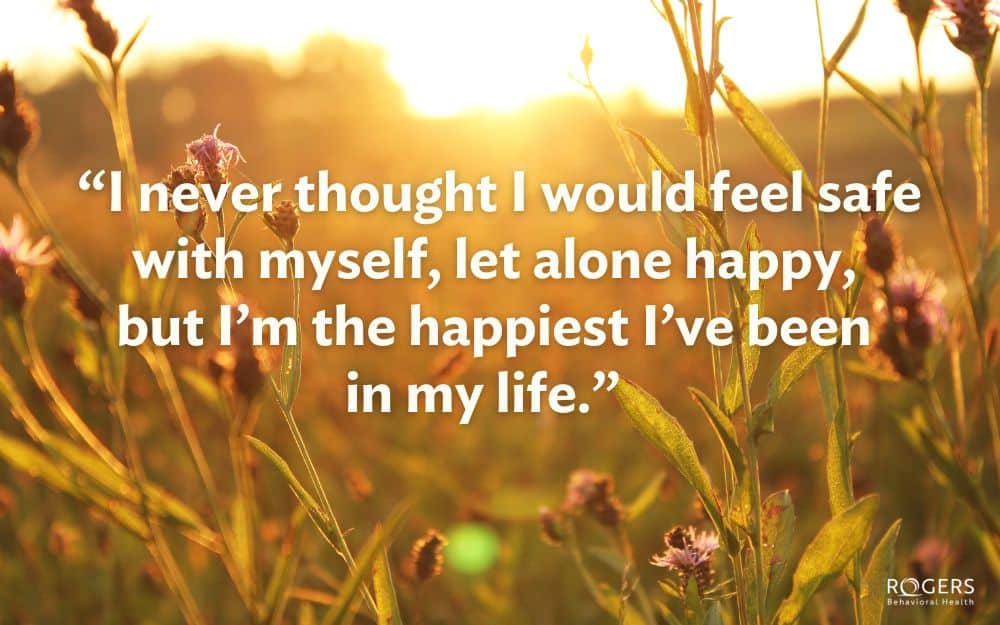 The image features a warm, sunlit field filled with tall grass and wildflowers. The sunlight creates a golden glow over the scene, with the sun setting or rising in the background. The tops of the grasses and flowers are highlighted by the light, casting soft shadows and creating a tranquil atmosphere. The image prominently features a quote written in bold white text across the center: “I never thought I would feel safe with myself, let alone happy, but I’m the happiest I’ve been in my life.”