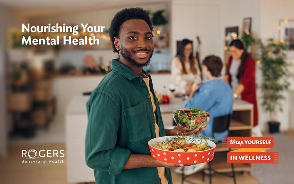 The image shows a smiling person in a green shirt holding a shallow red tray with white polka dots containing various foods, including greens and other vegetables. In their other hand, they hold a transparent bowl filled with a colorful salad. The background depicts a well-lit interior setting with a blurred group of people engaging in activities around a dining table, suggesting a social meal or gathering. There are warm, neutral tones throughout the room, contributing to a cozy atmosphere. A potted plant and some decorations are visible in the background. Text:Nourishing Your Mental Health Wrap Yourself In Wellness