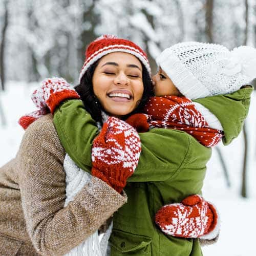 Two people in winter clothing hugging joyfully in a snowy forest.