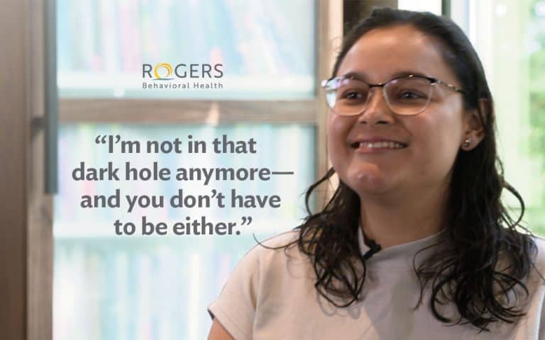 The image features a young woman who is a former Rogers OCD and anxiety patient. She has long dark hair and glasses and is smiling. A large quote is centered in the left half of the image: “I’m not in that dark hole anymore—and you don’t have to be either.”