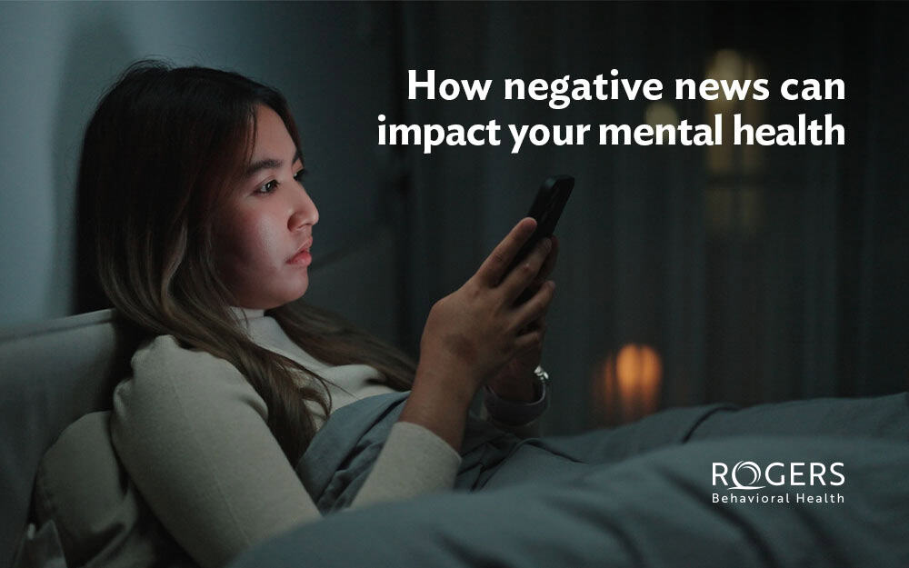 The image shows a woman sitting in a dimly lit room, intently looking at a smartphone looking at a smartphone. In large white text, is the phrase "How negative news can impact your mental health."