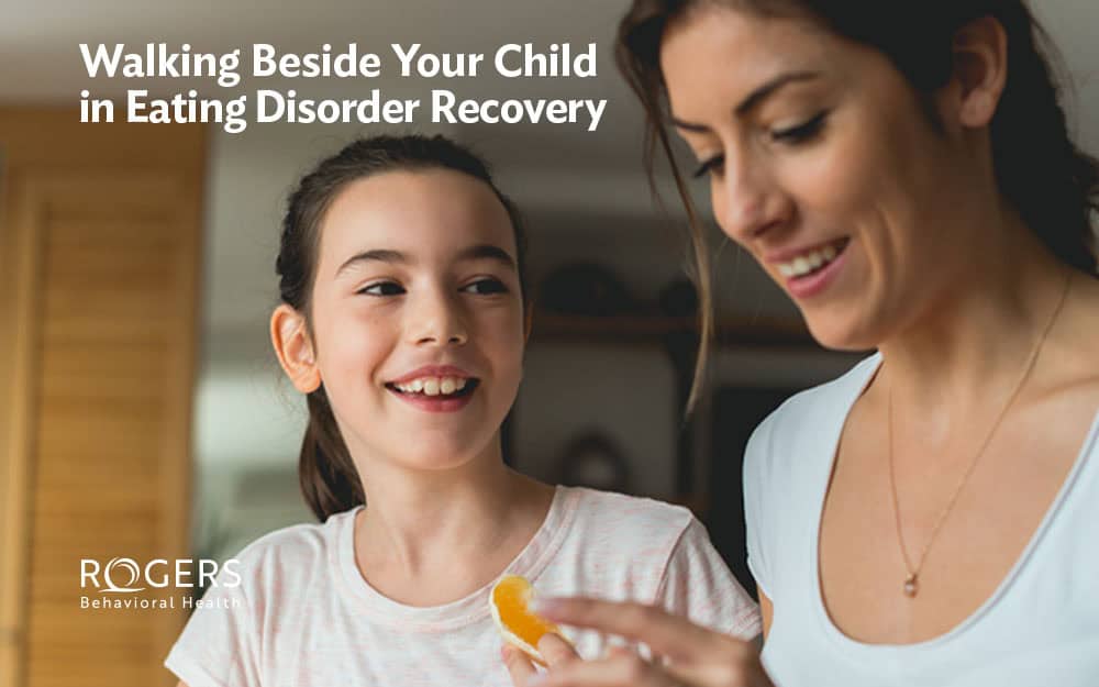 A mom and daughter home from eating disorder treatment, smiling together in a kitchen. The mom is prepping lunch and peeling an orange. Transcribed Text: Walking Beside Your Child in Eating Disorder Recovery