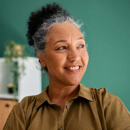 A smiling middle-aged woman with salt-and-pepper curly hair pulled back, wearing a brown button-up shirt, looking off to the side against a soft green background.