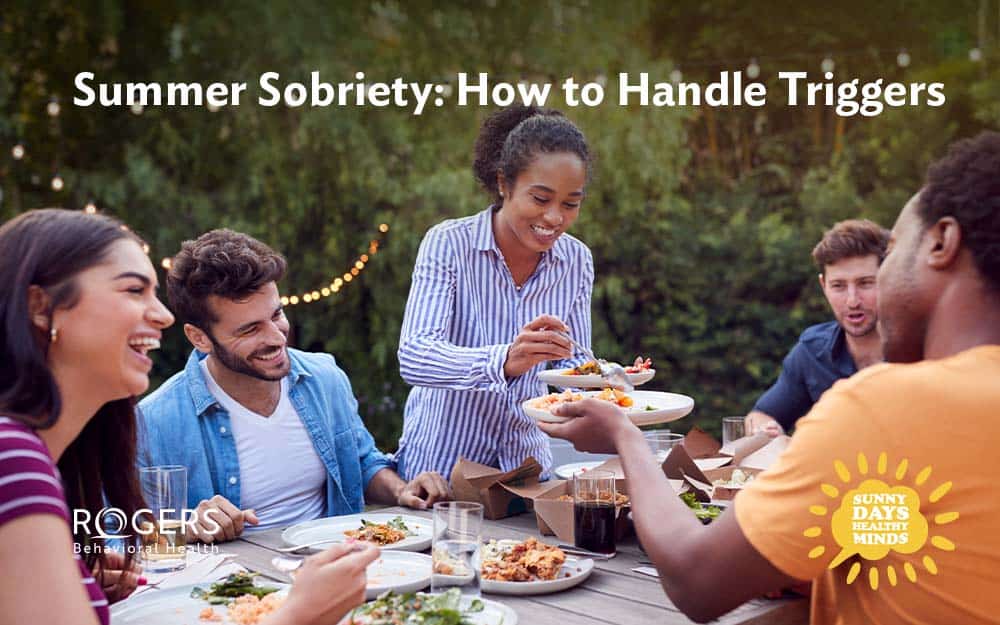 The image depicts a diverse group of people seated around a picnic table outdoors. They're talking and laughing, enjoying a summer meal without substances.