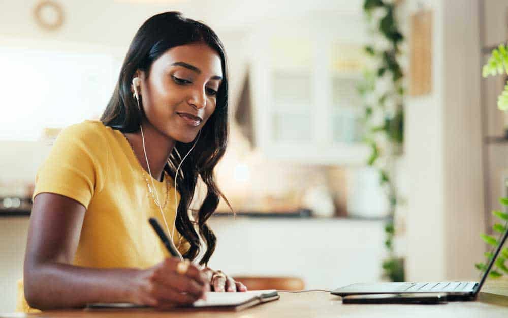 A woman takes notes while wearing earbuds and sitting in front of a computer.