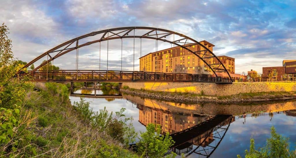 A modern arch bridge spans a tranquil river, reflecting its image on the water. In the background, a multi-story building glows in the setting sun, complemented by lush green foliage.