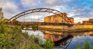 A modern arch bridge spans a tranquil river, reflecting its image on the water. In the background, a multi-story building glows in the setting sun, complemented by lush green foliage.