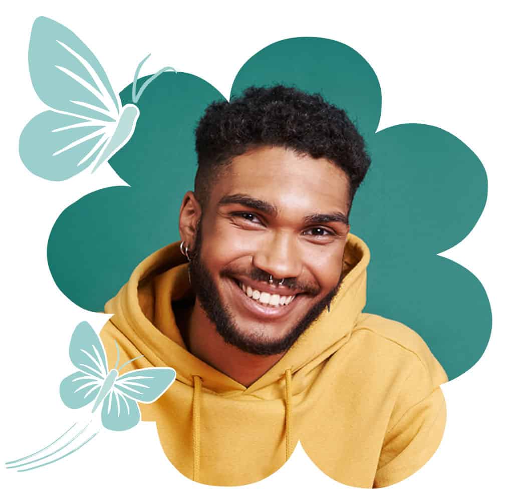 Portrait of a person wearing a yellow hoodie, shown within a teal flower-shaped graphic. Decorative light-blue butterfly illustrations appear in the upper left and lower left corners. The person has short curly hair, facial hair, and facial piercings, and is facing the camera.
