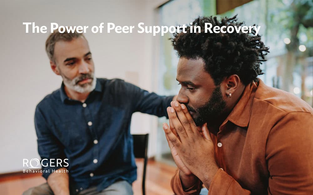 A recovery peer support specialist has his hand on the back of a man he's talking with in treatment. Whit text says: The Power of Peer Support In Recovery