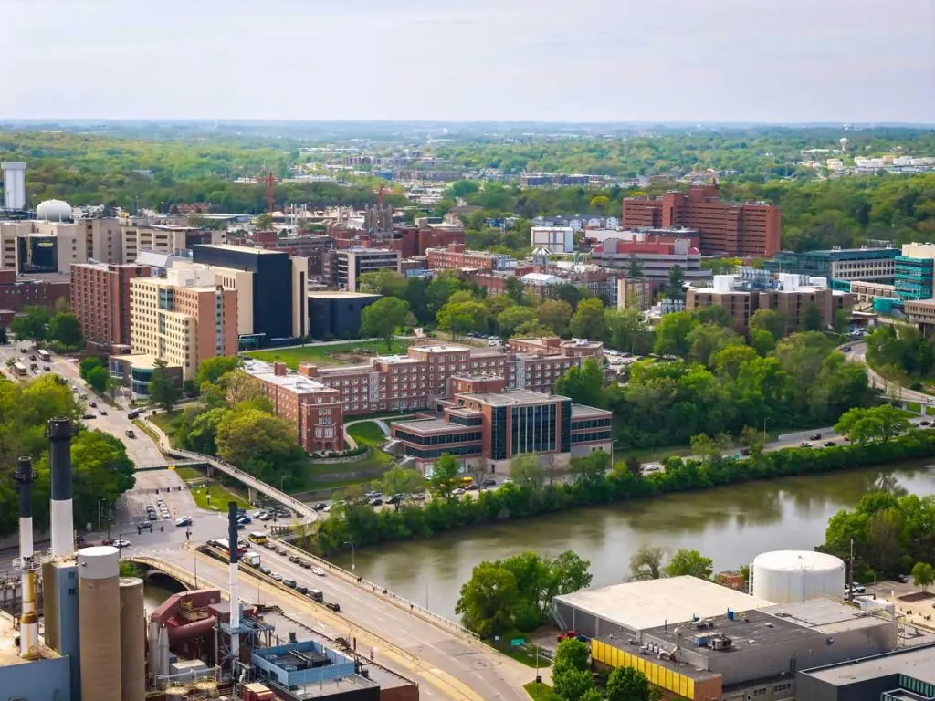 Aerial view of an urban area with large buildings surrounded by trees, a river running through the foreground, and a bridge carrying traffic across the water.