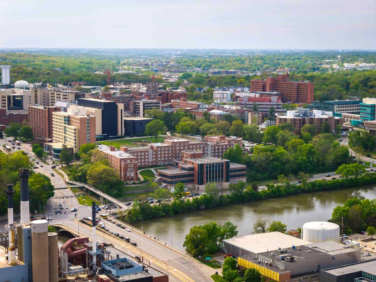 Aerial view of an urban area with large buildings surrounded by trees, a river running through the foreground, and a bridge carrying traffic across the water.