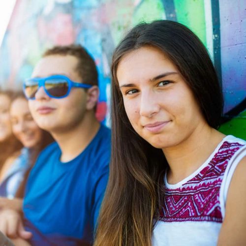 Young people sitting by a colorful graffiti wall, with a girl looking at the camera.